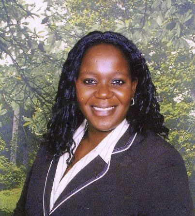 Portrait of a smiling woman with long black hair, wearing a dark blazer with white piping and a white blouse, outdoors with trees in the background.